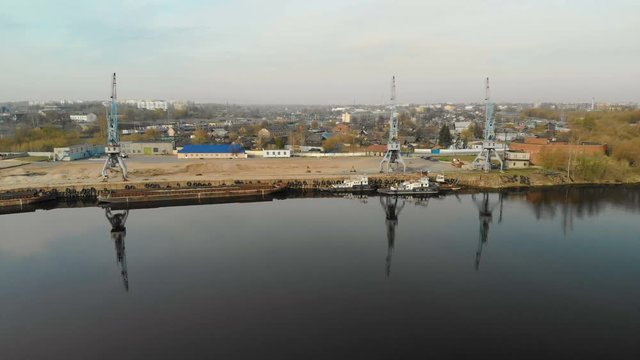 Aerial view of three cranes with reflection in the water in the river port with barges and tugs on the background of the city