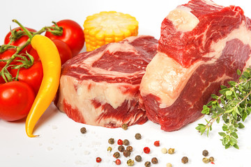 two fresh marbled beef steaks on a white plate with vegetables cherry tomatoes on a sprig, corn, spicy yellow pepper, a sprig of rosemary, salt and different varieties of peppers