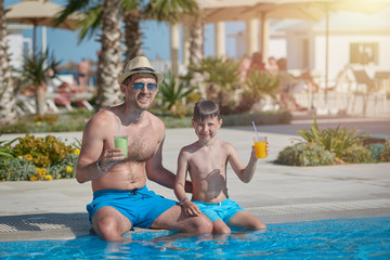 Fototapeta premium Dad and son enjoying their summer holidays. They are holding their cocktails in the hands, while sitting near the hotel’s swimming pool.