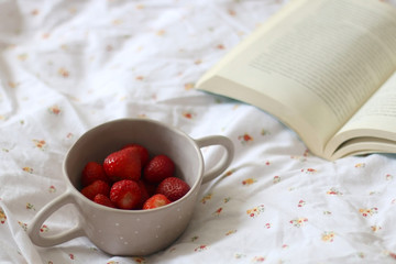 Bowl of strawberries and book on floral sheets. Leisure time concept. Selective focus.