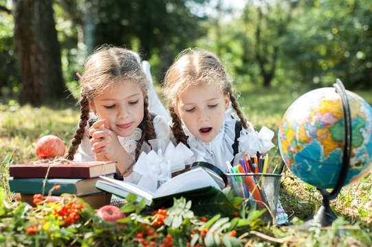 Schoolgirl girls are lying on the grass and reading books. Next to them is a globe, magnifying glass, pencils and an apple.