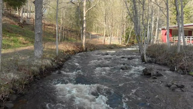 Drone footage flying really close to a fast flowing stream of white foamed water with trees and a building in the background. Filmed in realtime at 4K.