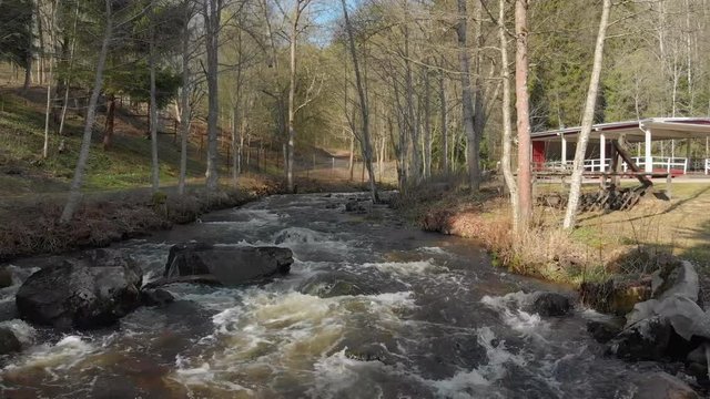 Drone footage flying really close to a fast flowing stream of white foamed water with some big rocks getting closer and a building in the background. Filmed in realtime at 4K.
