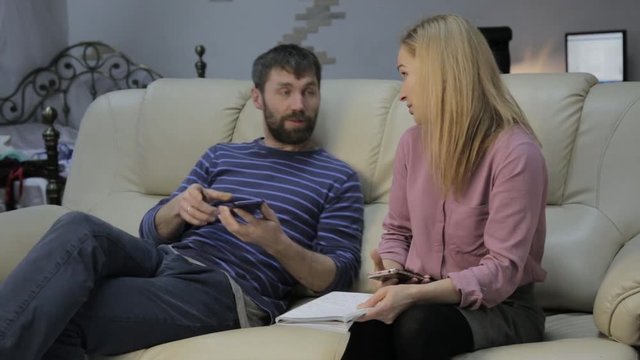 blonde student reads her notes, and her boyfriend sitting on sofa and uses smartphones