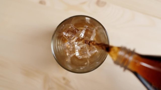 Top view of cola fizzy drink being poured in a glass with ice cubes on light brown wooden table, close up of cold sparkling soda pouring from bottle on beige background, slow motion