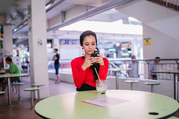 A young Malay Muslim woman is sitting at a hawker center and texting on her smartphone. She is young, elegant, beautiful and stylish and is wearing a hijab head scarf. 
