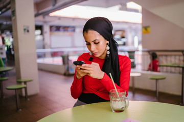 A young Malay Muslim woman is sitting at a hawker center and texting on her smartphone. She is...