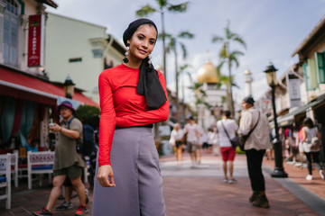 A beautiful, young, stylish and elegant Muslim Malay woman in a hijab head scarf on a street with a mosque in the day. She is smiling. She is about to go for Friday prayers.