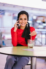 A girl in a red dress is talking to her boyfriend over the phone. She is enjoying her drink inside the cafe.