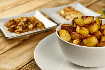 Roasted potatoes in a white bowl with rosemary and dill at next to fried onions and fish in batter on wooden background. Close-up rustic composition.