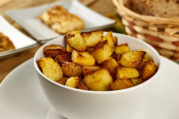Roasted potatoes in a white bowl with rosemary and dill at next to fried onions and fish in batter on wooden background. Close-up rustic composition.