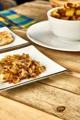 Roasted onions in a white bowl next to fried potatoes and cutlery on wooden background. Close-up rustic composition.