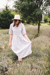 Stylish hipster girl in linen dress and hat walking in lavender field and relaxing. Happy bohemian woman enjoying lavender aroma in summer mountains. Atmospheric calm rural moment