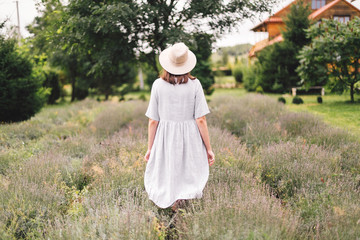 Stylish hipster girl in linen dress and hat walking in lavender field and relaxing. Happy bohemian...