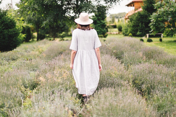 Stylish hipster girl in linen dress and hat walking in lavender field and relaxing. Happy bohemian...