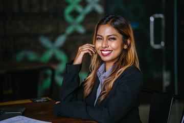 Business portrait of a juvenile Southeast Asian woman in a suit smiling broadly as she leans on a table in a trendy office room during the day.