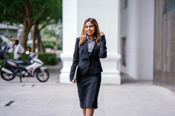 Fototapeta premium A beautiful and young Asian Malay woman is smiling confidently walking down a path by the street in the city during the day. She is professionally dressed and is holding her laptop.