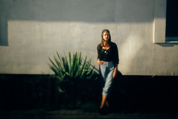 Stylish hipster girl posing in sunny light in street, atmospheric moment. Fashionable cool woman in denim jeans and black shirt standing at building with plant. Selective focus. Space text