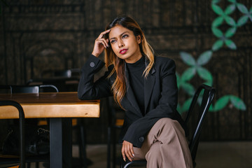  A young tanned professional Southeast Asian business woman in a turtle neck top looking away and is sitting on a bench in the city restaurant during the day.
