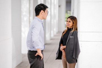 Candid shot of two diverse Asian business enthusiast, Korean man and a Malay woman having an animated conversation as they standby the hall during the day. They're both wearing a corporate attire.