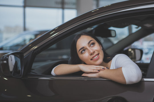 Happy Beautiful Dark Haired Woman Buying New Automobile At The Dealership, Sitting In A Car Looking Away Dreamily. Lovely Female Driver Daydreaming Of Adventures, Sitting In A New Auto, Copy Space