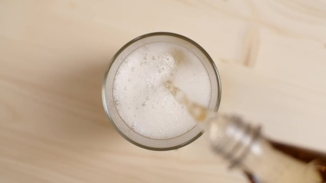 Top view of cream soda drink being poured in a glass with ice cubes on light brown wooden table, close up of cold fizzy sparkling soda pouring from bottle on beige background, slow motion