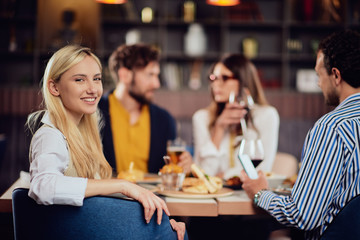 Young smiling Caucasian blonde cute woman dressed smart casual sitting at table in restaurant and looking over shoulder. In background are her friends having dinner.