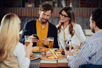 Young attractive Caucasian couple sitting in restaurant and looking at smart phone. In foreground their friends sitting adn drinking.