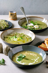 Variety of green vegetable soup asparagus, broccoli and pea, decorated by greens, vegetables, quail eggs, cream, olive oil, in ceramic bowls over grey spotted table with ingredients above.