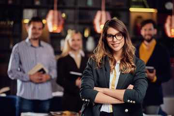 Smiling Caucasian businesswoman with long brown hait and dressed smart casual standing in restaurand with crossed arms. In background her successful team posing.