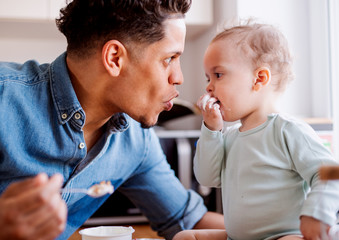 A father and a small toddler son eating fruit and yoghurt indoors at home.