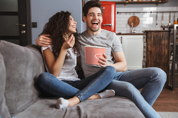 Picture of joyful young couple sitting on sofa at home and eating popcorn from bucket