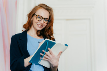 Prosperous satisfied businesswoman in elegant clothes writes in diary, has glad expression, wears spectacles, makes list of plans, writes text message in notebook, stands indoor. Female makes notes