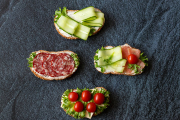Fresh sandwiches with sausage, cheese, bacon, tomatoes, lettuce, cucumbers on a dark background. Horizontal orientation