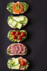 Fresh sandwiches with sausage, cheese, bacon, tomatoes, lettuce, cucumbers on a black background....