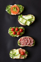 Fresh sandwiches with sausage, cheese, bacon, tomatoes, lettuce, cucumbers on a black background....