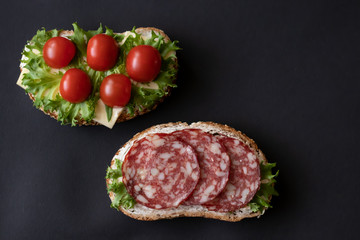 Fresh sandwiches with sausage, cheese, bacon, tomatoes, lettuce, cucumbers on a black background....