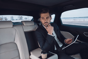 Fototapeta na wymiar Young perfectionist. Handsome young man in full suit working using digital tablet while sitting in the car
