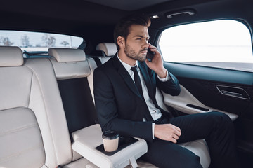 Business on the move. Handsome young man in full suit talking on the smart phone while sitting in the car