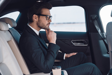 Thinking about solution. Thoughtful young man in full suit keeping hand on chin while sitting in the car