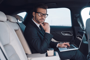 Fototapeta na wymiar Successful professional. Thoughtful young man in full suit working using laptop and adjusting his eyewear while sitting in the car