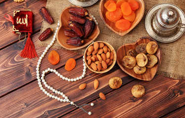 Ramadan concept. Delicious dates, dried figs, dried apricots, walnuts, almonds, raisins on bamboo and silver plates, rosary beads with Islamic holy book Quran on a pine wooden table background.