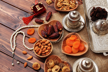 Ramadan concept. Delicious dates, dried figs, dried apricots, walnuts, almonds, raisins on bamboo and silver plates, rosary beads with Islamic holy book Quran on a pine wooden table background.