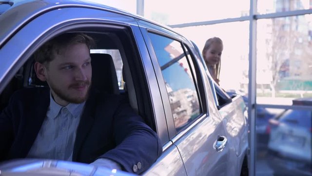 happy family in car dealership, little funny girl into trunk of automobile is played hide and seek with dad in cabine while buying new vehicle at showroom
