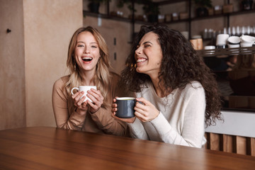 Happy girls friends sitting in cafe talking with each other drinking tea or coffee.