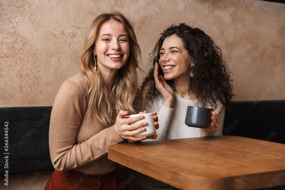 Poster Happy girls friends sitting in cafe talking with each other drinking tea or coffee.