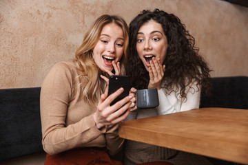 Emotional shocked girls friends sitting in cafe using mobile phone.