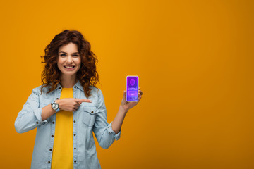 cheerful curly redhead woman pointing with finger at smartphone with e shopping app on screen on orange