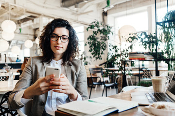 Portrait of beautiful business lady sitting and working at the city eco cafe with mobile phone in hands. Notepad, laptop, coffee, desert on the table