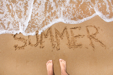 Top view of female feet standing on the beach sand with wave motion coming.Word summer writting on the sand.Summer travel and relax concept.